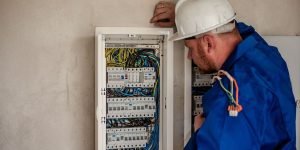 A professional electrician wearing a hard hat and blue protective clothing inspects a breaker panel with multiple wires and circuit breakers, ensuring safety during the home renovation.