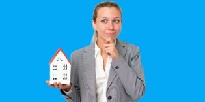 A woman in a gray blazer holds a model house with a thoughtful expression against a blue background, considering tenanted properties in your area.