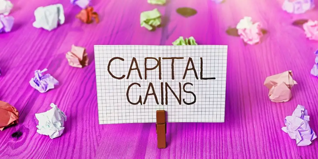 A sign with "CAPITAL GAINS" scrawled on graph paper is clipped to a table, surrounded by crumpled pieces of paper on a pink wooden surface, evoking the chaotic journey of navigating tax complexities.