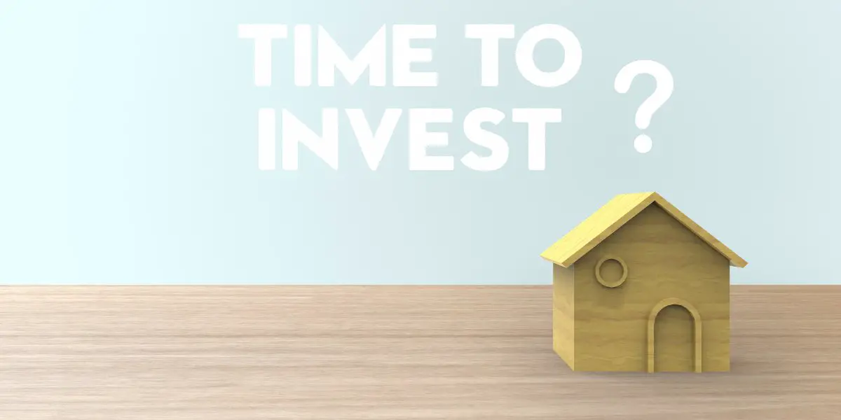 A small wooden house model sits on a wooden surface with the words "TIME TO INVEST?" displayed in bold white text on a light blue background, hinting that it might be the perfect time to invest in rental property.