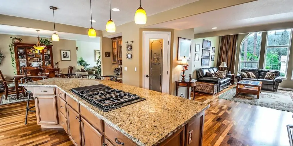 Modern open plan kitchen and living area featuring a granite island, gas stovetop, and wooden cabinetry. The space is complete with a dining table, leather sofas, large windows, and hardwood floors, reflecting the latest in kitchen design trends.