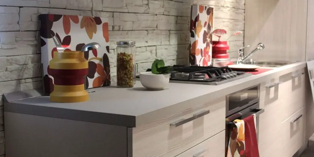 Discover modern kitchen design with a light countertop, a sleek gas stove, and captivating artwork featuring abstract leaf designs. A jar of pasta, bowl, and vibrant yellow red vase adorn the counter against a textured wall backdrop, creating an elegant culinary space.