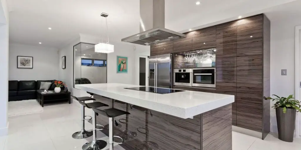 Discover the latest in kitchen trends with this modern kitchen, featuring a long island, bar stools, and dark wood cabinetry. Built in ovens sit beneath a stylish chandelier. A cozy seating area with a sofa and wall art completes this chic design.