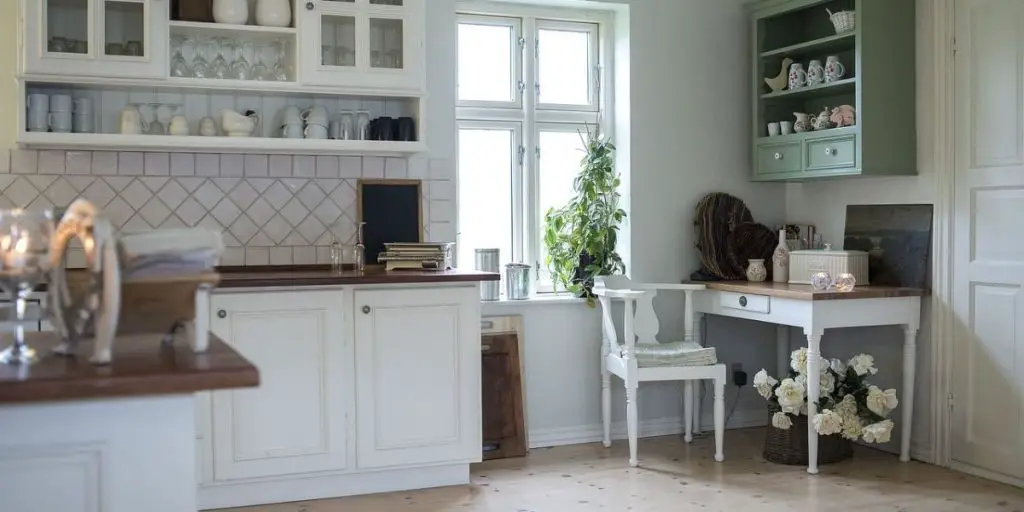 Cozy kitchen with white cabinets, wooden countertops, and a small desk. A window with a plant offers natural light. Green and white dishes add charm to the modern kitchen design, complemented by a vase of flowers on the floor.