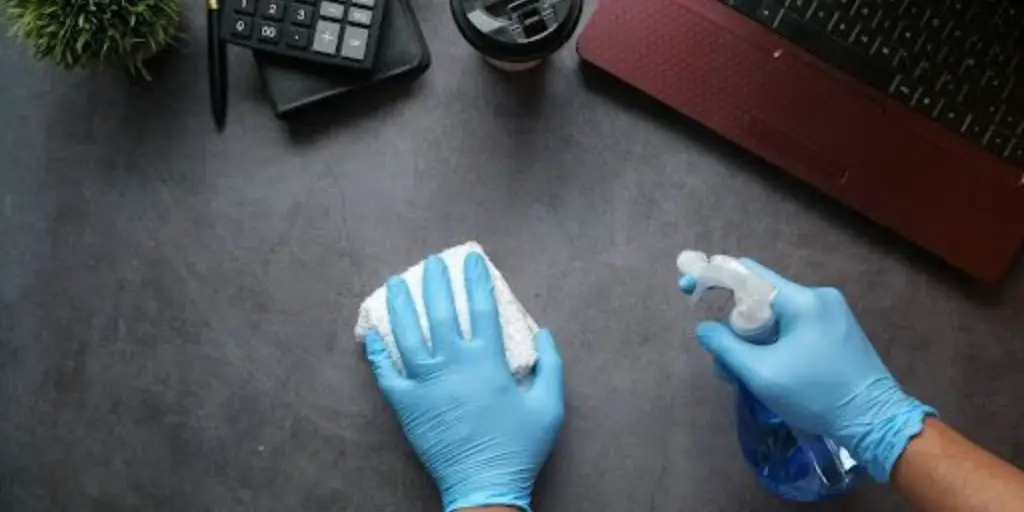 Person wearing blue gloves wipes a desk with a sponge and cleaning spray. Nearby are a laptop, a calculator, a pen, a plant, and a coffee cup.