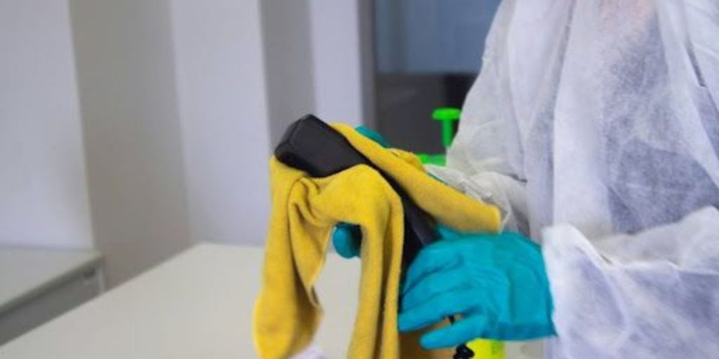 Person wearing protective gloves and suit cleaning a phone handset with a yellow cloth on a white table.