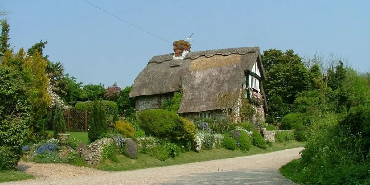 A quaint cottage with Old World Charm, featuring a thatched roof and surrounded by greenery. The stone walled structure sits beside a gravel path, reminiscent of Devonshire's idyllic scenery, and is beautifully landscaped with various bushes and plants.