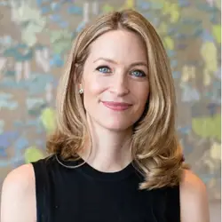 A woman with shoulder length blonde hair, wearing a sleeveless black top, stands in front of a blurred, multicolored background.
