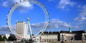 The London Eye Ferris wheel on the South Bank of the River Thames, with nearby buildings under a blue sky with scattered clouds.